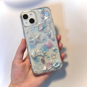Puede incluir: Funda transparente para teléfono decorada con conchas marinas, perlas y una estrella de mar. La funda tiene un diseño transparente con detalles en azul, rosa y blanco. La funda está diseñada para un smartphone.