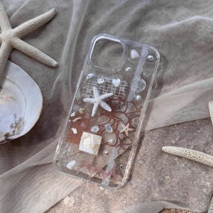 May include: A clear phone case adorned with seashells, starfish, and coral elements. The case incorporates various white and beige seashells, small white starfish, and coral-like features. The design includes a square mother-of-pearl element and small white pearls.