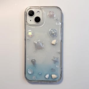 May include: Clear phone case with a gradient blue and white background, adorned with sea-themed embellishments. Features a stingray, starfish, shells, and pearl accents. The case is designed to fit a smartphone.