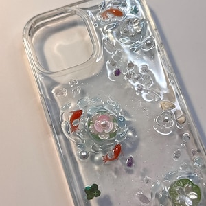 Può includere: Una custodia per telefono trasparente con decorazioni tridimensionali incorporate, tra cui pesci koi arancioni, fiori di loto rosa e bianchi, piccole perle bianche, foglie di ninfea verdi e minuscole pietre viola e trasparenti in resina.