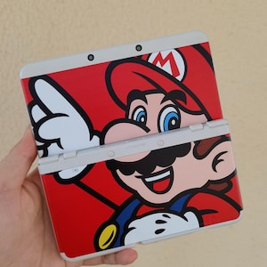 Pode incluir: Uma consola de jogos portátil com um exterior vermelho vibrante e um gráfico grande da personagem Mario. O dispositivo é apresentado na posição aberta, exibindo o icónico boné vermelho, as jardineiras azuis e a luva branca da personagem.