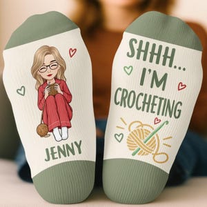 May include: A pair of off-white socks with sage green cuffs and toes. One sock features an illustration of a person with glasses crocheting, the name JENNY, and small red hearts. The other sock displays the text SHHH... I'M CROCHETING.