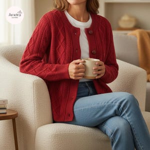 Puede incluir: Un cárdigan rojo de punto trenzado con botones de madera. El suéter de manga larga tiene un cuello redondo y se usa sobre una camiseta blanca. La persona está sentada en una silla color crema y sosteniendo una taza beige.