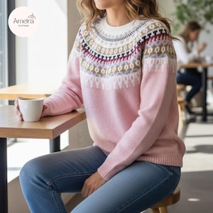 May include: A pink sweater with a patterned yoke in white, navy, burgundy, and gold. The sweater is worn with blue jeans. A white coffee cup rests on a wooden table. The Ameira Clothing logo is in the upper left corner.