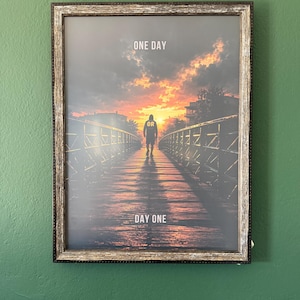 May include: A framed wall art print featuring a silhouette of a person walking on a bridge toward a sunset. The text ONE DAY is at the top, OR is on the person's back, and DAY ONE is at the bottom. The frame has a weathered, dark brown finish.