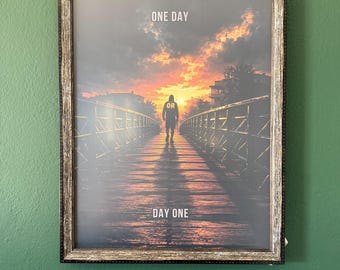 May include: A framed wall art print featuring a silhouette of a person walking on a bridge toward a sunset. The text ONE DAY is at the top, OR is on the person's back, and DAY ONE is at the bottom. The frame has a weathered, dark brown finish.
