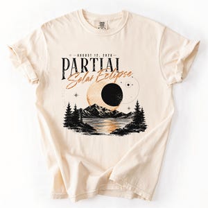 May include: A cream-colored short-sleeve t-shirt featuring a graphic of a partial solar eclipse over a mountain lake. The text reads "AUGUST 12, 2026" and "PARTIAL Solar Eclipse" in black and orange script, surrounded by pine trees and stars.