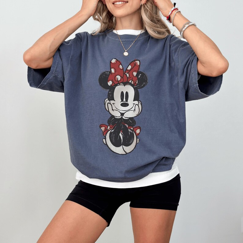 May include: Blue-grey oversized t-shirt featuring a Minnie Mouse graphic. The graphic shows Minnie Mouse with a red polka-dot bow, black ears, and a red skirt. The shirt has short sleeves and a white undershirt peeking out from the neckline.