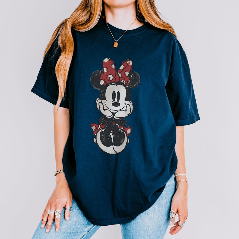 May include: Navy blue t-shirt featuring a distressed graphic of Minnie Mouse. Minnie is depicted with a red bow with white polka dots. The shirt is a relaxed fit, suitable for casual wear. The design is centered on the front of the shirt.