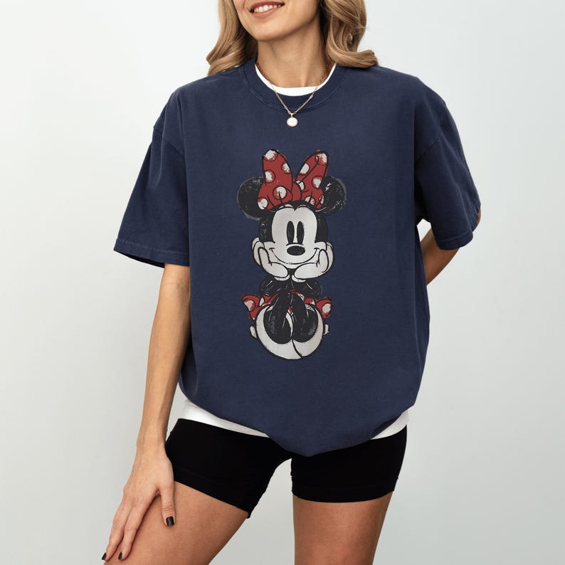 May include: Navy blue t-shirt featuring a distressed Minnie Mouse graphic with a red polka dot bow. The shirt has a relaxed fit and a white undershirt peeking from the neckline. The overall style is casual and comfortable.
