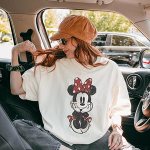 May include: Cream-colored t-shirt featuring a distressed Minnie Mouse graphic with a red bow and polka dots. The cartoon character is in a seated pose. The shirt is worn by a person in a car, wearing a corduroy hat and sunglasses.