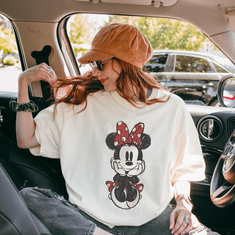 May include: Cream-colored t-shirt featuring a distressed Minnie Mouse graphic with a red bow and polka dots. The cartoon character is in a seated pose. The shirt is worn by a person in a car, wearing a corduroy hat and sunglasses.