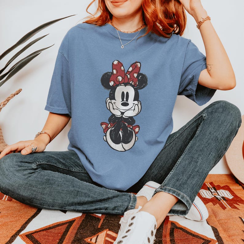 May include: A light blue t-shirt featuring a Minnie Mouse graphic. The cartoon character is depicted in a seated pose with a red polka dot bow. The shirt is made of a soft material and has short sleeves.