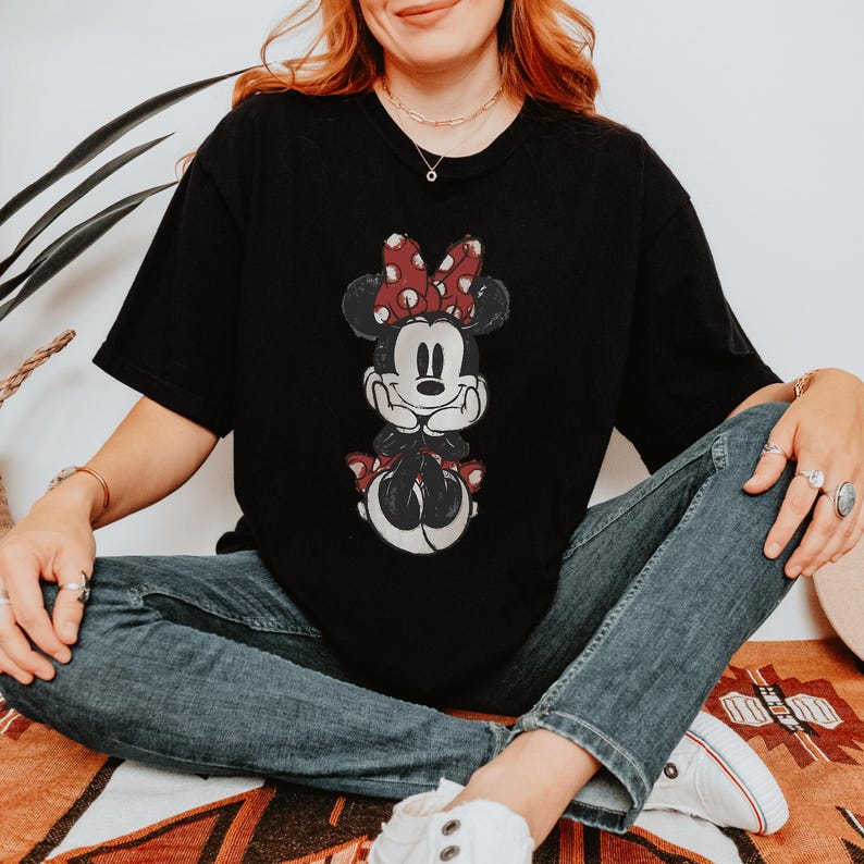 May include: Black t-shirt featuring a graphic of Minnie Mouse. The cartoon character is depicted with a red and white polka dot bow, black ears, and a smiling face. The shirt is a classic crew neck style.