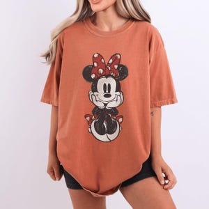 May include: Burnt orange t-shirt featuring a graphic of Minnie Mouse. The cartoon character has a red bow with white polka dots. The shirt has short sleeves and a relaxed fit, perfect for casual wear.