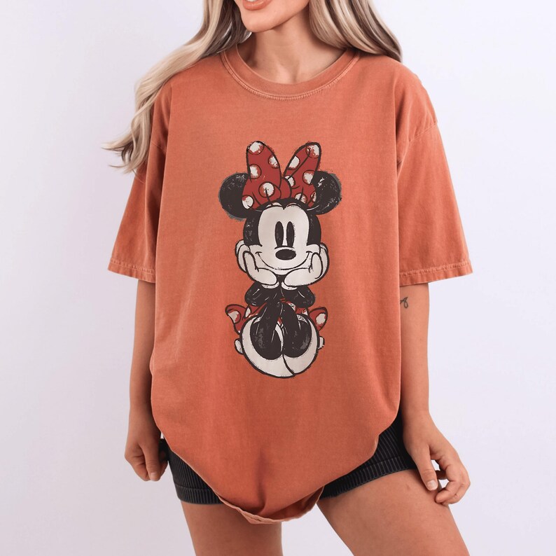 May include: Burnt orange t-shirt featuring a graphic of Minnie Mouse. The cartoon character has a red bow with white polka dots. The shirt has short sleeves and a relaxed fit, perfect for casual wear.