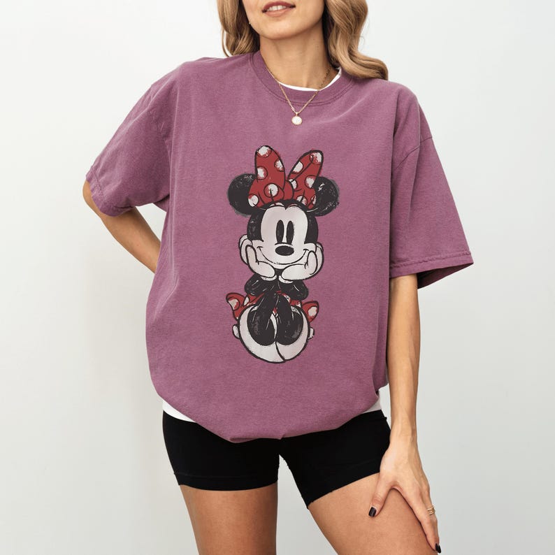 May include: A mauve-colored t-shirt featuring a graphic of Minnie Mouse. The cartoon character is depicted with a red bow with white polka dots, black ears, and a black and white outfit. The shirt has short sleeves and a crew neck.