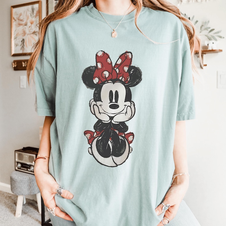 May include: A light blue t-shirt featuring a distressed graphic of Minnie Mouse. Minnie is depicted with a red polka dot bow, black ears, and a red skirt. The shirt has short sleeves and a relaxed fit.