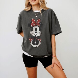 May include: Dark gray t-shirt featuring a distressed graphic of Minnie Mouse with a red polka dot bow. The shirt has short sleeves and a relaxed fit. The shirt is paired with black shorts.