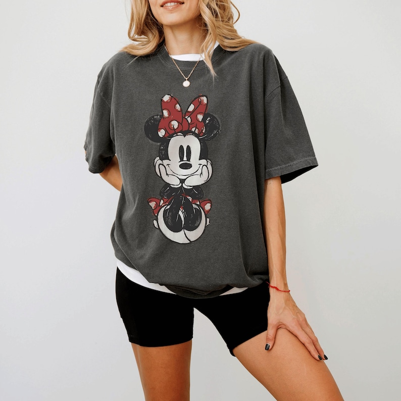 May include: Dark gray t-shirt featuring a distressed graphic of Minnie Mouse with a red polka dot bow. The shirt has short sleeves and a relaxed fit. The shirt is paired with black shorts.