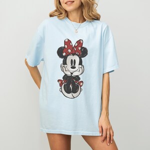 May include: Light blue t-shirt featuring a Minnie Mouse graphic. The graphic shows Minnie Mouse with a red polka dot bow and red polka dot accents. The shirt is a casual, oversized fit.