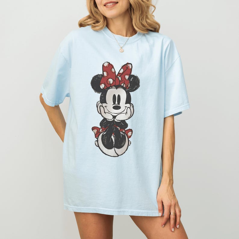 May include: Light blue t-shirt featuring a Minnie Mouse graphic. The graphic shows Minnie Mouse with a red polka dot bow and red polka dot accents. The shirt is a casual, oversized fit.