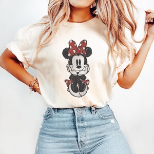May include: Cream-colored t-shirt featuring a Minnie Mouse graphic. The cartoon character is depicted in black, white, and red, with a polka-dot bow. The shirt is paired with blue denim jeans.