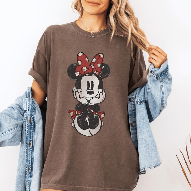 May include: A brown t-shirt featuring a distressed graphic of Minnie Mouse. Minnie Mouse is depicted with a red bow with white polka dots and a matching red skirt. The shirt has short sleeves and a relaxed fit.