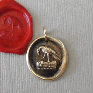 May include: A gold-colored pendant with a bird design, next to a red wax seal. The pendant features a bird perched on a log. The wax seal has a similar bird design. The background is a light gray color.