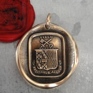 Puede incluir: Un colgante dorado con un diseño de escudo detallado, con un escudo, llaves cruzadas y la palabra "YIELD". Un sello de cera rojo es visible en la esquina superior izquierda. El colgante tiene un lazo para una cadena.
