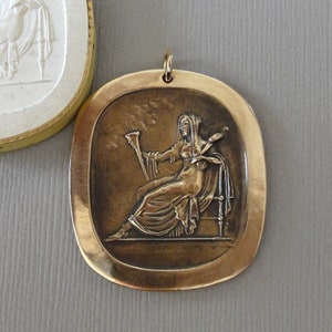 May include: A bronze pendant featuring a raised relief of a woman seated on a throne, holding a staff and a scroll. The pendant is oval shaped with a smooth, slightly textured surface.