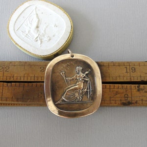 May include: A bronze pendant featuring a raised relief of a woman holding a musical instrument. The pendant is oval shaped and has a smooth, slightly textured surface.