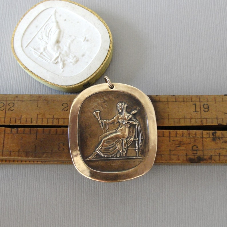 May include: A bronze pendant featuring a raised relief of a woman holding a musical instrument. The pendant is oval shaped and has a smooth, slightly textured surface.