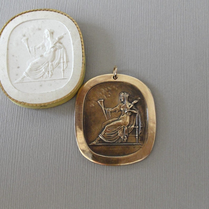 May include: A bronze pendant featuring a raised relief of a woman seated on a throne, holding a staff and a scroll. The pendant is oval shaped with a smooth, slightly textured surface.