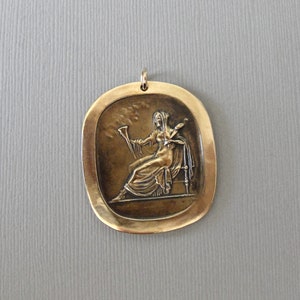 May include: A bronze pendant featuring a relief sculpture of a woman holding a horn and a sword. The pendant is oval shaped and has a textured surface.
