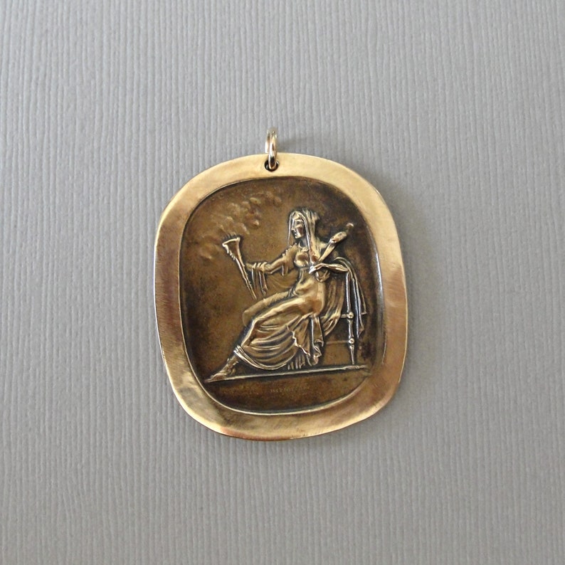 May include: A bronze pendant featuring a relief sculpture of a woman holding a horn and a sword. The pendant is oval shaped and has a textured surface.