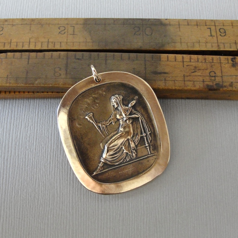 May include: A bronze pendant featuring a raised relief image of a woman holding a paintbrush and palette. The pendant is oval shaped with a smooth, slightly textured surface.