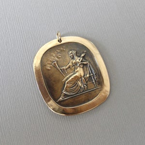 May include: A gold-toned pendant featuring a raised relief sculpture of a woman seated on a throne, holding a staff and a spindle. The pendant is oval-shaped with a smooth, slightly textured surface.