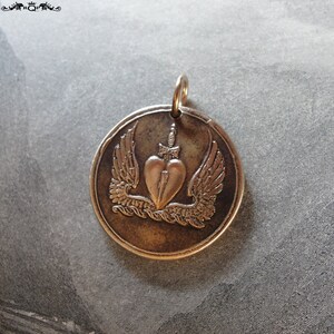 May include: A silver pendant with a heart pierced by a dagger and surrounded by wings. The pendant is on a gray background.