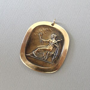 May include: A gold-toned pendant featuring a raised relief image of a woman holding a torch and sitting on a throne. The pendant is oval-shaped and has a smooth, polished surface.