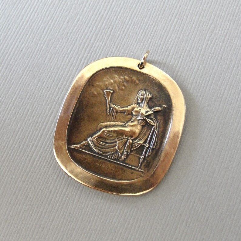 May include: A gold-toned pendant featuring a raised relief image of a woman holding a torch and sitting on a throne. The pendant is oval-shaped and has a smooth, polished surface.