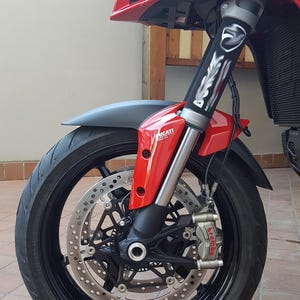 May include: Close-up of a red and black motorcycle front end. The image highlights the front wheel, brake discs, and suspension components. The wheel is black with a tire, and the brake calipers are branded "Brembo". The motorcycle is a Ducati.