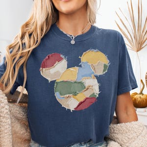 May include: A navy blue t-shirt featuring a patchwork Mickey Mouse design in various colors. The shirt has a crew neck and short sleeves. The word "denim" is printed at the bottom right corner of the image.