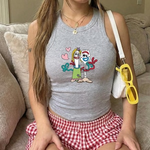 May include: A heather gray ribbed tank top featuring a graphic print of two cartoon characters with googly eyes, one wearing a green dress and the other with red pipe cleaner arms, surrounded by small pink hearts. The top has a crew neckline.