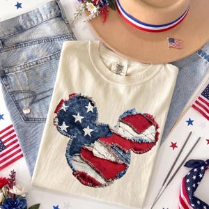May include: Cream-colored t-shirt with a patriotic Mickey Mouse design, light-wash jeans, a tan hat with red, white, and blue trim, and American flag accessories. The shirt features a patchwork design of the American flag.