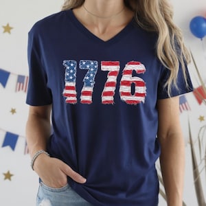 May include: Navy blue V-neck t-shirt with the number "1776" in a distressed American flag design. The shirt is a patriotic apparel item, perfect for Independence Day or other national holidays. The shirt is made of a soft material.