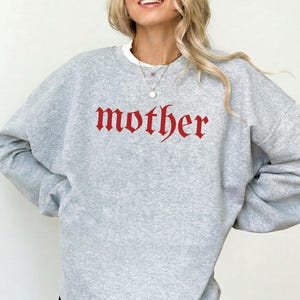 May include: An athletic heather gray crewneck sweatshirt featuring the word mother printed in a red gothic-style font across the chest. The garment has long sleeves, a ribbed neckline, and a relaxed fit, paired with black shorts.