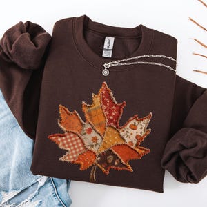 May include: A dark chocolate brown crewneck sweatshirt featuring a large, central maple leaf applique made from various fabric patches with frayed edges. The patches include orange and white gingham, plaid, floral, and pumpkin patterns.
