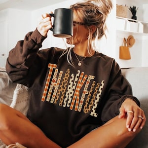 May include: A dark brown long-sleeve crewneck sweatshirt featuring the word THANKFUL printed across the chest in large block letters. Each letter displays a unique pattern, including orange plaid, floral motifs, leopard print, and polka dots.