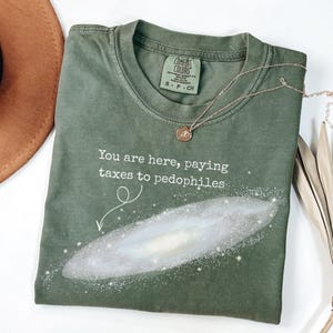 Puede incluir: Camiseta verde oliva con el texto "You are here, paying taxes to pedophiles" y un gr&aacute;fico de galaxia. Un collar dorado con un colgante est&aacute; sobre la camiseta. La camiseta es de la marca Comfort Colors.